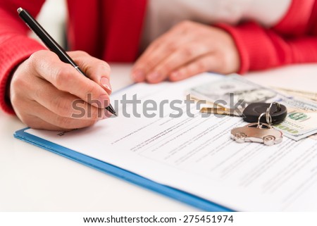 Closeup of woman auto dealer signing rental contract in the office. Car key and money on papers. Shallow depth of field.