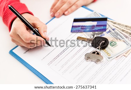 Young female leasing consultant signing car insurance contract in the office. Closeup of sold vehicle key with credit card and cash. Shallow depth of field.