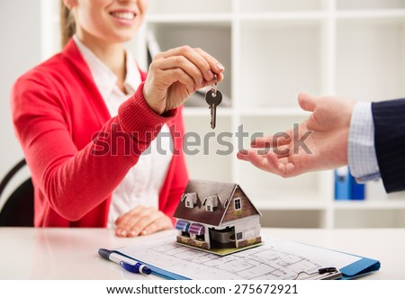 Closeup of smiling woman realtor giving key of new apartment to client. Female house agent sitting at the desk with house miniature and plan.