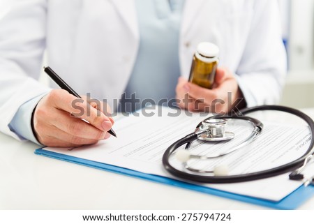 Female doctor filling medical form on clipboard holding ballpoint and medicine bottle. Healthcare and insurance concept.