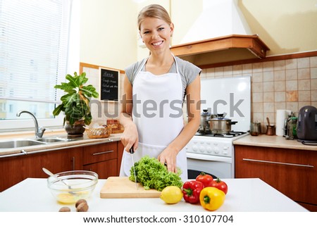 Happy woman cutting salad preparing healthy breakfast. Young female in white apron cooking meal for her family.