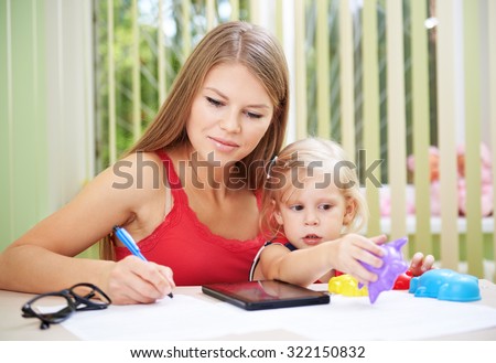 Young busy female telecommuting while her child is playing with toys. Pretty mom with cute daughter checking family bills and documents.