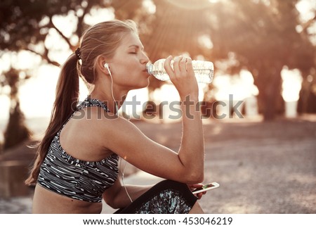 Thirsty female jogger drinking fresh water and listening music in headphones after training. Young athletic woman exercising in the park outdoors.