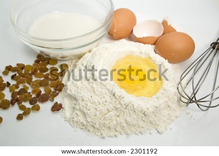 An egg in a pile of flour preparing for baking with kitchen tools sugar and raisins