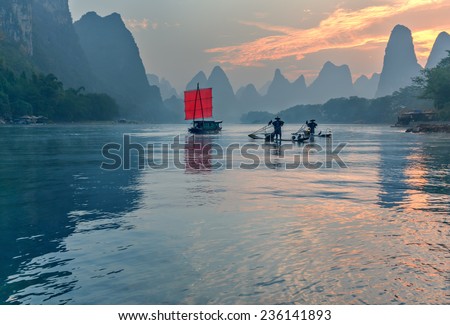 XINGPING, CHINA - OCTOBER 22, 2014: Fisherman stands on traditional bamboo boats at sunrise (boat with a red sail in the background) - The Li River, Xingping, China