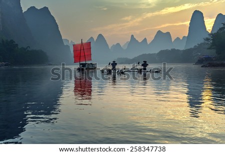 XINGPING, CHINA - OCTOBER 22, 2014: Fishermans stands on traditional bamboo boats at sunrise (boat with a red sail in the background) - The Li River, Xingping, China