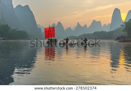 Fisherman stands on traditional bamboo boats at sunrise (boat with a red sail in the background) - The Li River, Xingping, China