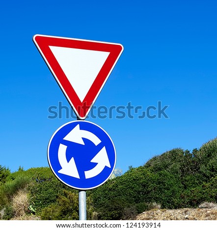 roundabout and give away signs surrounded by vegetation under a blue sky