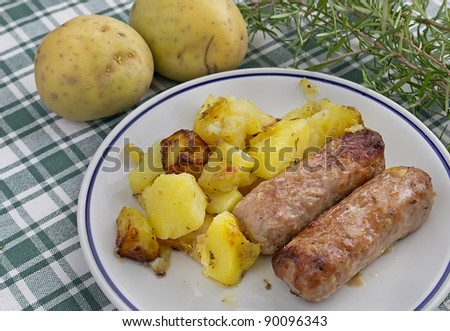 sausages and cooked potatoes in a plate surrounded by rosemary and raw potatoes on a checkered table cloth