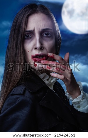 Scary female vampire with a blood on her lip. Night sky with the full moon in the background.