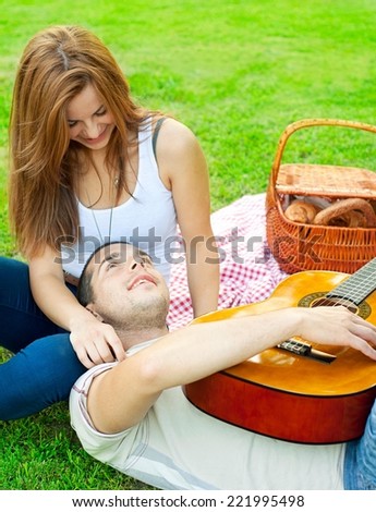 Young couple lying in the park on a summer day. Picnic basket in the background. Man is playing a guitar.