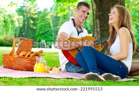 Young couple lying in the park on a summer day. Picnic basket in the background. Man is playing a guitar.