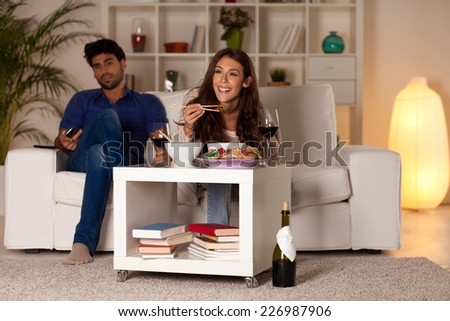 Couple sitting in the living room, watching TV. Woman eating.