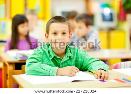 Cute little boy sitting in the classroom and smiling. Elementary age. Other kids in the background. NOTE: All the drawings and artwork in the classroom are made by children.