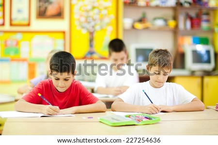 Two cute little boys sitting in the classroom and writing. Elementary age. Other kids in the background. NOTE: All the drawings and artwork in the classroom are made by children.