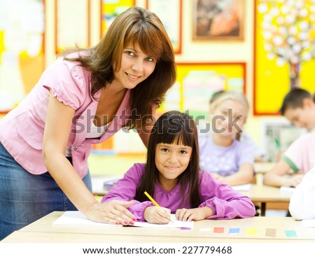 Beautiful female teacher explaining something to a little girl sitting in the classroom. Elementary age.  NOTE: All the drawings and artwork in the classroom are made by children.