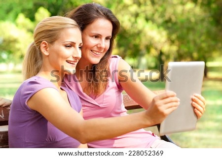 Two friends in the park sitting on a bench and taking pictures with tablet computer.