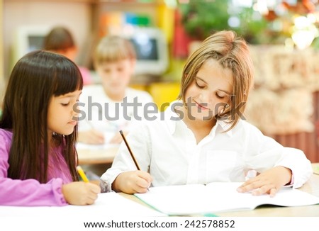 Cute little girls sitting in the classroom and writing. Elementary age. Other kids in the background.