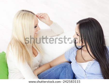 Two sisters sitting on the sofa at home and talking. Blonde and dark haired girl.