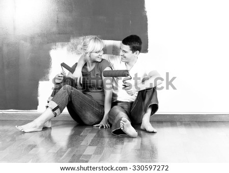 Young caucasian couple sitting on the floor and smiling in front of partially painted wall - black and white, intentionally added grain