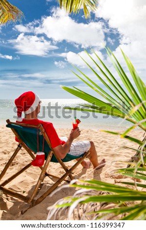 Tropical Christmas. Man in Santa\'s hat with cocktail sitting on chair on a beach under palm trees.