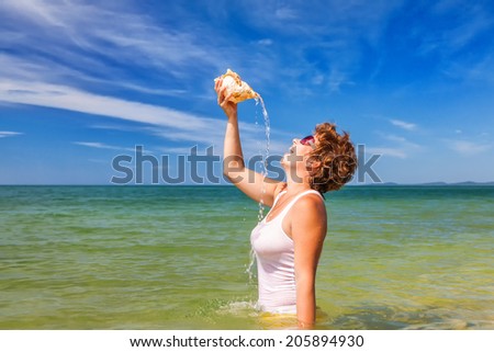 Beach holiday. A smiling brown hair woman staying at water and pours water from a large conch shell onto herself
