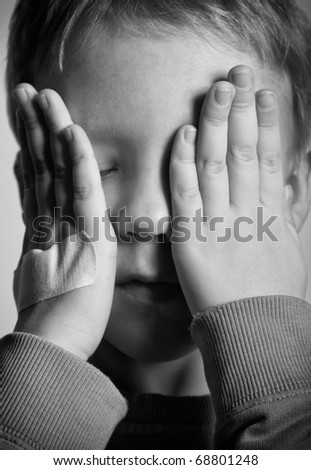 BW portrait of sad crying little boy covers his face with hands. One hand with medical plaster.