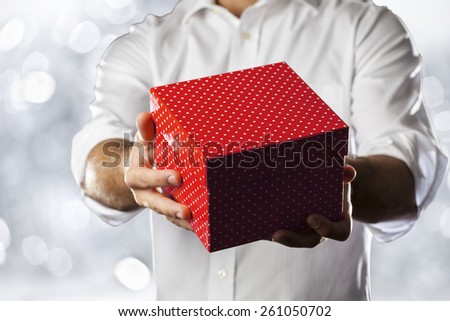 A Man holding a gift box in a gesture of giving in a silver lights background.