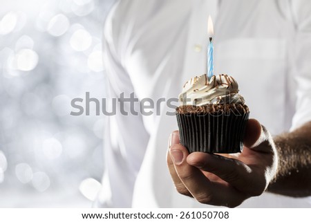 A Man holding a cupcake with a candle in a silver lights background.