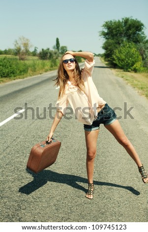 Attractive young woman with suitcase hitchhiking along a road.
