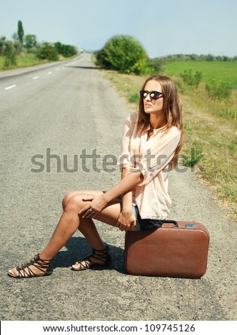 Beautiful girl in black glasses sitting on a suitcase by a countryside road