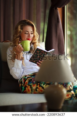 Happy young woman drinking coffee and reading book on couch near the window at home