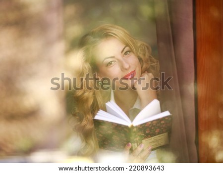 Smiling woman with book at home, view through window glass with reflections and flares