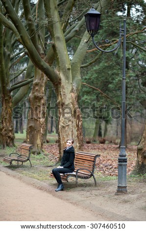 Happy young woman in a black coat sitting on a wooden bench on the background of autumn park