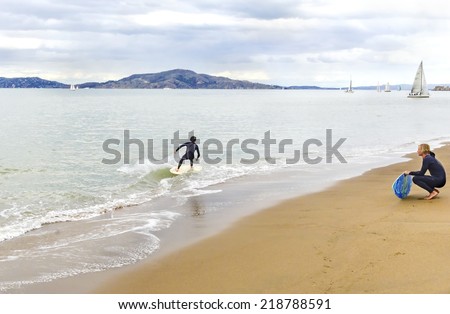 SAN FRANCISCO,USA - MARCH 1 2014: View of surfers in wet suits surfing his board or skimboarding in shallow water in the bay beach coast,sailboats and yachts crossing the sea in Marina, California.
