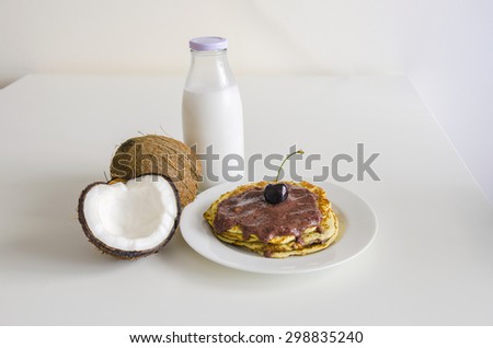 Breakfast and brunch coconut pancakes meal with cracked open coco, glass bottle of milk and cherry on white background. Stack of crepes is topped with cream syrup ready to eat.