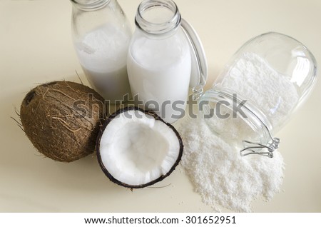 Coconut products. Cracked open coconut with meat cut in half, grounded flakes in a mason jar, flour and fresh milk in glass bottles on a table shot from the top.