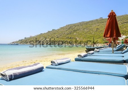 Sun beds with cushions, umbrellas, golden sand and the blue sea in Mykonos, Greece. A greek island summer holiday scene at the Psarou beach with a boat in the crystal clear water over a bushy hill.