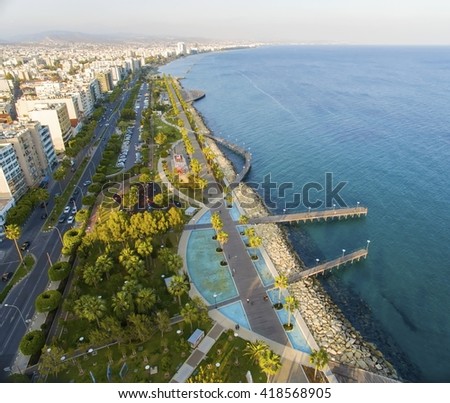Aerial view of Molos Promenade on the coast of Limassol city in Cyprus. A view of the walk path surrounded by palm trees, pools of water, grass, the Mediterranean sea, piers, rocks and urban skyline.