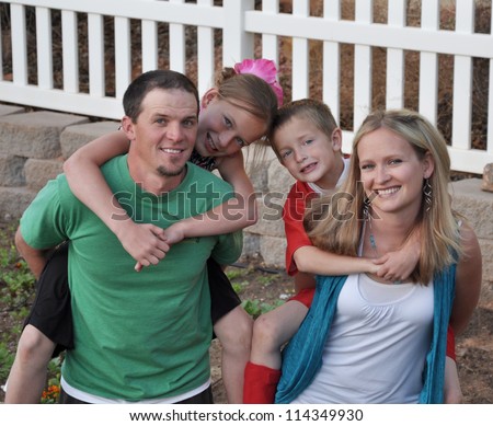 A cute young family doing piggy back rides in their picket fenced back yard.