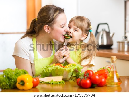 Mother Feeding Kid Vegetables In Kitchen