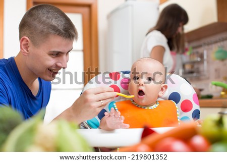 young dad feeding his baby and mom cooking at kitchen
