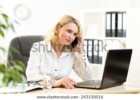 Middle-aged businesswoman working in office. Business lady talking on mobile phone and looking at laptop