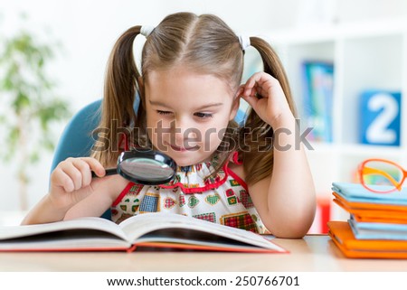 Funny child girl reads book using magnifier while sitting at table at home