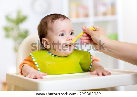 Mother feeding baby girl. Child sitting in hich chair in kitchen.