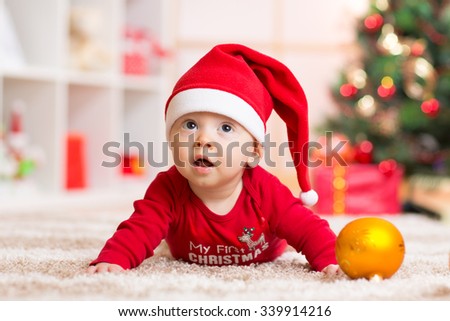 Happy Smiling baby lying against domestic festive backdrop with Christmas tree. Cute kid wearing Christmas Santa hat and suit