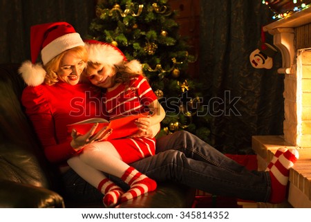 Middle-aged mother and her child daughter reading a book by a Christmas tree in cozy living room in winter