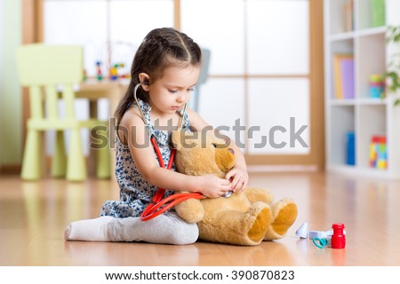 Little child girl with stethoscope and teddy bear sitting on floor, on home interior background