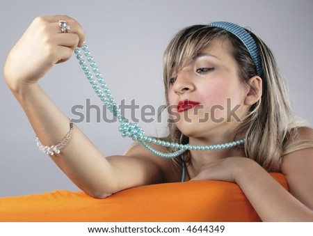 Blond Pin-up style fascinating girl with light blue shirt seat on orange sofa, playing wiht light blue pearl necklace.