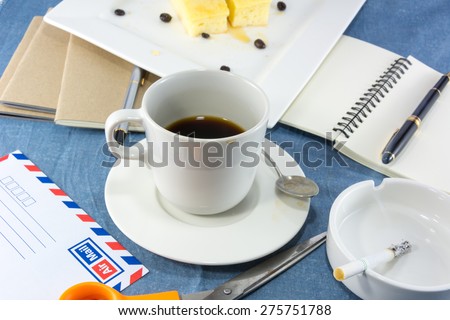 Clutter workspace with cup of coffee, blank note and pen, butter cake on white plate, letter envelope with scissors and cigarette in ashtray